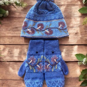 May include: Blue knitted winter hat with a pom pom and matching mittens. Both hat and mittens have embroidered floral designs.
