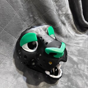 Puede incluir: M&aacute;scara de animal de piel negra y blanca con detalles verdes alrededor de los ojos y la nariz. La m&aacute;scara tiene un dise&ntilde;o detallado con agujeros en toda la superficie.