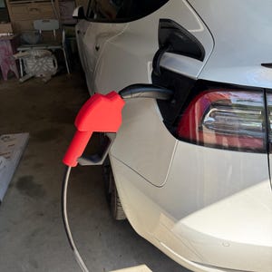 May include: A white electric car is plugged in to charge. The charging cable is black with a red handle. The car is parked in a garage.