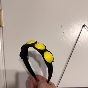 Op de afbeelding: Zwarte haarband met drie grote, heldergele cirkelvormige accenten. De haarband wordt in een hand gehouden tegen een witte achtergrond. De gele accenten zijn gelijkmatig verdeeld over de bovenkant van de haarband.