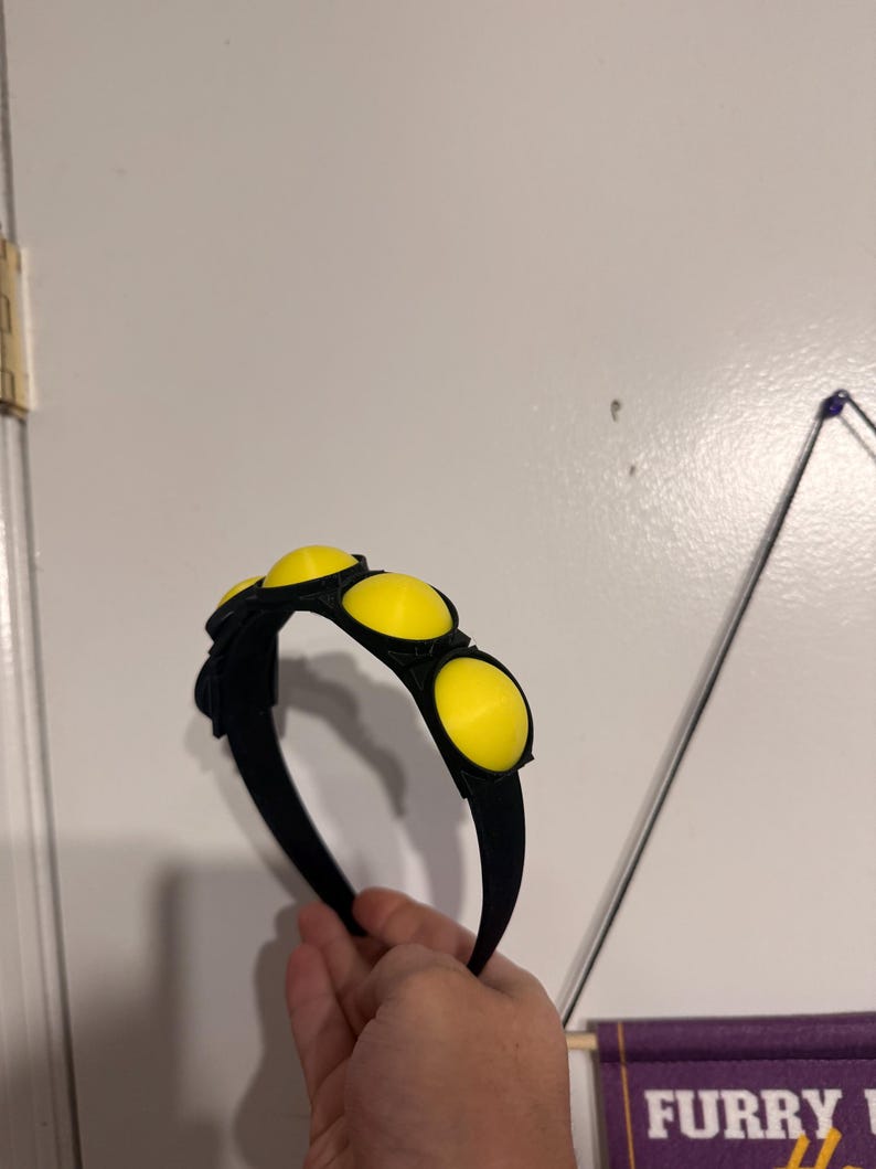 Op de afbeelding: Zwarte haarband met drie grote, heldergele cirkelvormige accenten. De haarband wordt in een hand gehouden tegen een witte achtergrond. De gele accenten zijn gelijkmatig verdeeld over de bovenkant van de haarband.