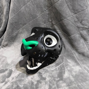 Puede incluir: M&aacute;scara de animal de peluche negro y blanco con nariz verde y ojos blancos. La m&aacute;scara est&aacute; hecha de un material ligero y tiene un dise&ntilde;o detallado.