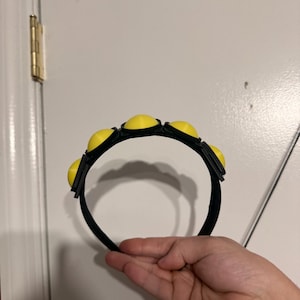 Op de afbeelding: Een zwarte haarband met zes gelijkmatig verdeelde, felgele, afgeronde versieringen. De haarband wordt door een hand vastgehouden, waardoor het ontwerp en het kleurcontrast worden benadrukt. Het accessoire is waarschijnlijk voor mode- of kostuumdoeleinden.