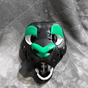 Puede incluir: M&aacute;scara de animal de peluche negra y verde con boca y nariz blancas. La m&aacute;scara tiene un aspecto realista y est&aacute; hecha de un material suave y afelpado.
