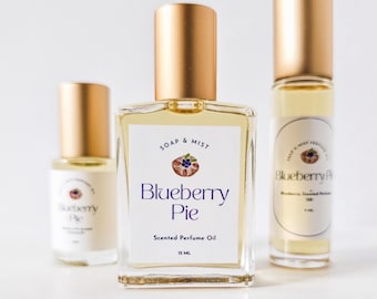 May include: Three glass perfume bottles with gold caps. The central bottle is rectangular and labeled "Blueberry Pie" with "Scented Perfume Oil" and "15 ML". The other two bottles are smaller, with similar labels. The bottles are filled with a light yellow liquid.