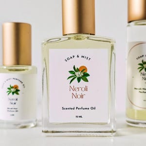 May include: Three perfume oil bottles with gold lids. The central bottle has a white label with "SOAP & MIST" and "Neroli Noir" with a botanical illustration. The label also states "Scented Perfume Oil" and "15 ml".