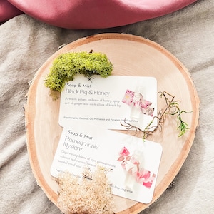 Op de afbeelding: Twee kleine witte kaartjes met roze bloemenpatronen en tekst die "Soap & Mist Black Fig & Honey" en "Soap & Mist Pomegranate Mysiere" luidt. De kaartjes liggen op een houten oppervlak met groen mos.
