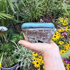 Könnte beinhalten: Ein kleiner, handgehaltener Keramikübertopf mit einem strukturierten, braunen Körper mit Blumenmuster und einem kontrastierenden, glatten, blaugrünen Rand. Das Gefäß hat eine breite, offene Oberseite und einen stabilen Boden.