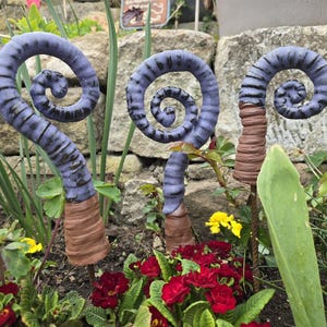 Könnte beinhalten: Drei dekorative Gartenstecker mit einem Spiraldesign. Die Stecker haben eine lila und schwarz gestreifte Spiralspitze und eine braune, strukturierte Basis. Sie sind in einem Garten mit roten Blumen und grünem Laub platziert.