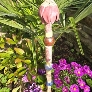 Può includere: Un paletto da giardino decorativo con una parte superiore in ceramica rosa e bianca a forma di fiore. Il paletto ha un fusto a motivi con perline di legno e ceramica. Il paletto è circondato da piante verdi e fiori viola.