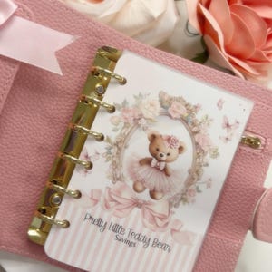 May include: A pink planner with gold rings, holding an insert with a teddy bear illustration. The insert reads "Pretty Little Teddy Bear Savings" and is decorated with flowers and bows. Peach roses are in the background.