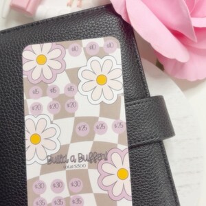 May include: A planner insert with a daisy and checkerboard design. The insert has amounts from £5 to £40, with the text "Build a Buffer!" and "total £500". The insert is on a black planner with a pink flower and accessories.