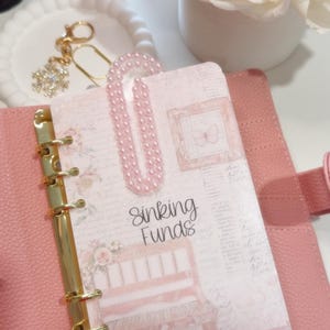 May include: A pink planner with gold rings, open to a page labelled "Sinking Funds." The page has a floral design and a pink pearl paper clip. A white floral arrangement and a decorative tray with a charm are in the background.