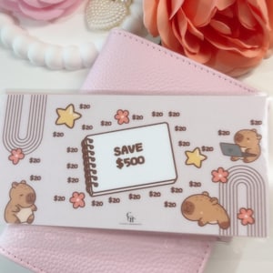 Puede incluir: Un sobre de presupuesto rosa con un diseño que presenta ilustraciones de estrellas, flores y carpinchos. El sobre tiene el texto "SAVE 500$" y está rodeado de cantidades de 20$. El sobre está encima de una cartera rosa.