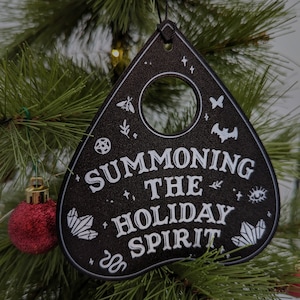 May include: Black triangular ornament with a white circle in the center and the text "Summoning the Holiday Spirit" with various symbols around the text. The ornament is hanging on a green Christmas tree.