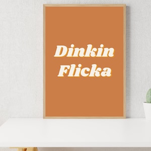 May include: A framed print with an orange background and white text that reads "Dinkin Flicka".