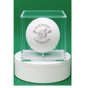 May include: A white lacrosse ball with the text "McKinney Lacrosse" and a logo, displayed inside a clear acrylic case. The case sits on a white, round base against a green background.