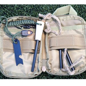 K&ouml;nnte beinhalten: Ge&ouml;ffnetes, beigefarbenes Survival-Kit mit verschiedenen Werkzeugen. Es enth&auml;lt einen Feuerstarter mit wei&szlig;em Griff, ein blaues Multi-Tool und mehrere schwarze St&auml;be. Das Kit ist ge&ouml;ffnet und zeigt den Inhalt vor einem gr&uuml;nen Hintergrund.
