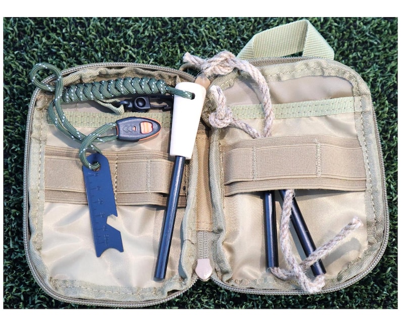 K&ouml;nnte beinhalten: Ge&ouml;ffnetes, beigefarbenes Survival-Kit mit verschiedenen Werkzeugen. Es enth&auml;lt einen Feuerstarter mit wei&szlig;em Griff, ein blaues Multi-Tool und mehrere schwarze St&auml;be. Das Kit ist ge&ouml;ffnet und zeigt den Inhalt vor einem gr&uuml;nen Hintergrund.