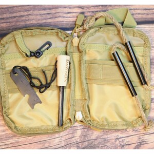 K&ouml;nnte beinhalten: Ge&ouml;ffnete, beigefarbene Survival-Kit-Tasche mit Rei&szlig;verschluss. Sie enth&auml;lt einen schwarzen Karabiner, ein Metall-Multitool, einen Feuerstarter mit "DAWSON"-Aufdruck und zwei schwarze Metallst&auml;be, die an einem Seil befestigt sind.