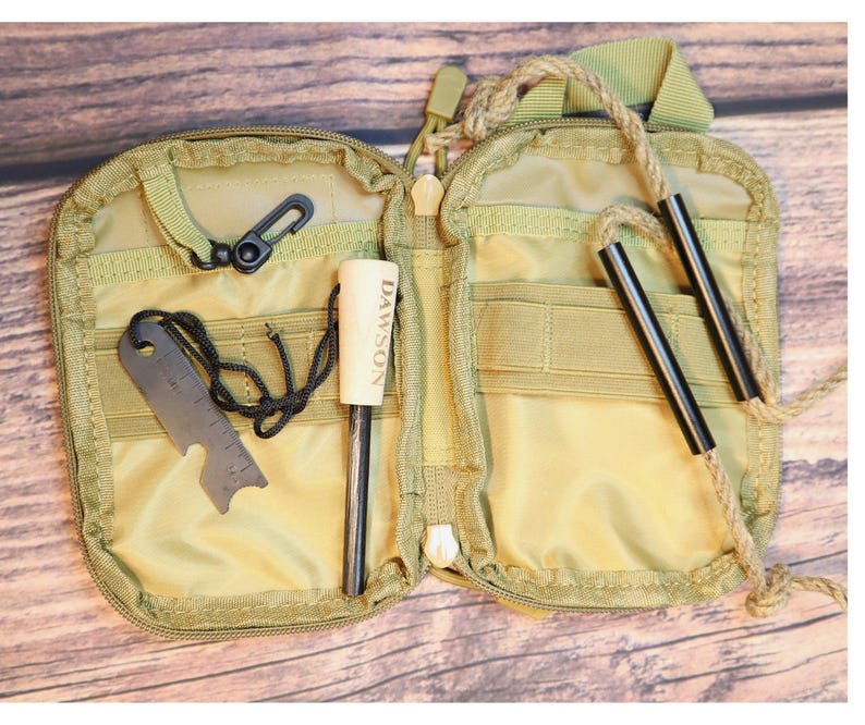 K&ouml;nnte beinhalten: Ge&ouml;ffnete, beigefarbene Survival-Kit-Tasche mit Rei&szlig;verschluss. Sie enth&auml;lt einen schwarzen Karabiner, ein Metall-Multitool, einen Feuerstarter mit "DAWSON"-Aufdruck und zwei schwarze Metallst&auml;be, die an einem Seil befestigt sind.
