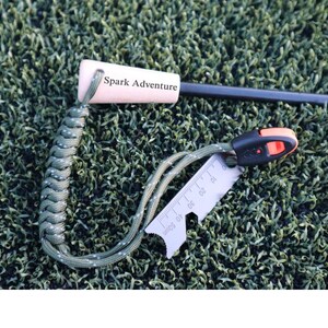 K&ouml;nnte beinhalten: Ein Survival-Feuerstarter-Kit mit Holzgriff, schwarzem Ferrostab und einem kleinen Multitool mit Lineal. Der Griff tr&auml;gt den Text "Spark Adventure". Das Kit ist an einem gr&uuml;n-wei&szlig;en geflochtenen Lanyard befestigt.