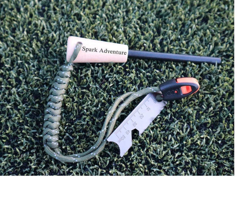 K&ouml;nnte beinhalten: Ein Survival-Feuerstarter-Kit mit Holzgriff, schwarzem Ferrostab und einem kleinen Multitool mit Lineal. Der Griff tr&auml;gt den Text "Spark Adventure". Das Kit ist an einem gr&uuml;n-wei&szlig;en geflochtenen Lanyard befestigt.