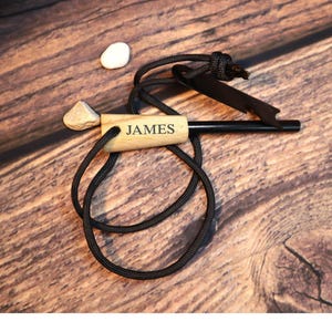 K&ouml;nnte beinhalten: Ein Holzfeuerstarter mit dem eingravierten Namen "JAMES". Er ist an einer schwarzen Schnur befestigt und enth&auml;lt einen schwarzen Metallz&uuml;nder. Zwei kleine Steine sind ebenfalls vorhanden.