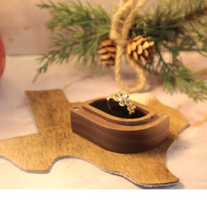 May include: A gold ring with a floral design sits in a wooden ring box shaped like the state of Texas. The box is on a brown, fuzzy surface.