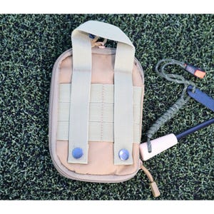 K&ouml;nnte beinhalten: Beige Nylon-Tasche mit Griff und zwei vertikalen Riemen, mit Rei&szlig;verschluss und zwei blauen Druckkn&ouml;pfen. Ein Survival-Tool-Kit enth&auml;lt einen schwarzen Feuerstarter, einen wei&szlig;en Griff und eine gr&uuml;ne Kordel.