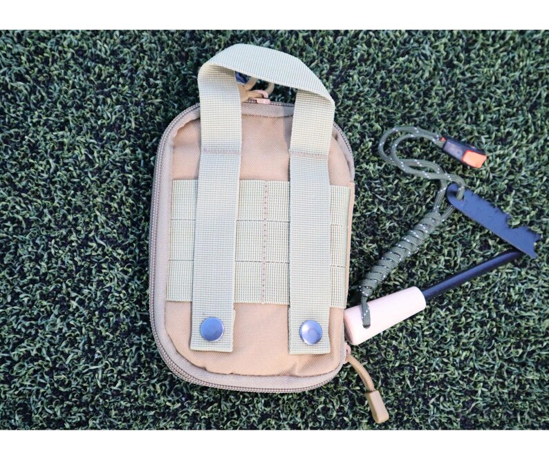 K&ouml;nnte beinhalten: Beige Nylon-Tasche mit Griff und zwei vertikalen Riemen, mit Rei&szlig;verschluss und zwei blauen Druckkn&ouml;pfen. Ein Survival-Tool-Kit enth&auml;lt einen schwarzen Feuerstarter, einen wei&szlig;en Griff und eine gr&uuml;ne Kordel.