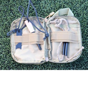 K&ouml;nnte beinhalten: Ge&ouml;ffnete, beigefarbene Survival-Kit-Tasche mit verschiedenen Werkzeugen. Enth&auml;lt einen schwarzen Feuerstarter, zwei Ferro-St&auml;be, einen Holzanz&uuml;nder und eine Schnur. Die Tasche ist ge&ouml;ffnet und zeigt den Inhalt vor einem gr&uuml;nen Hintergrund.
