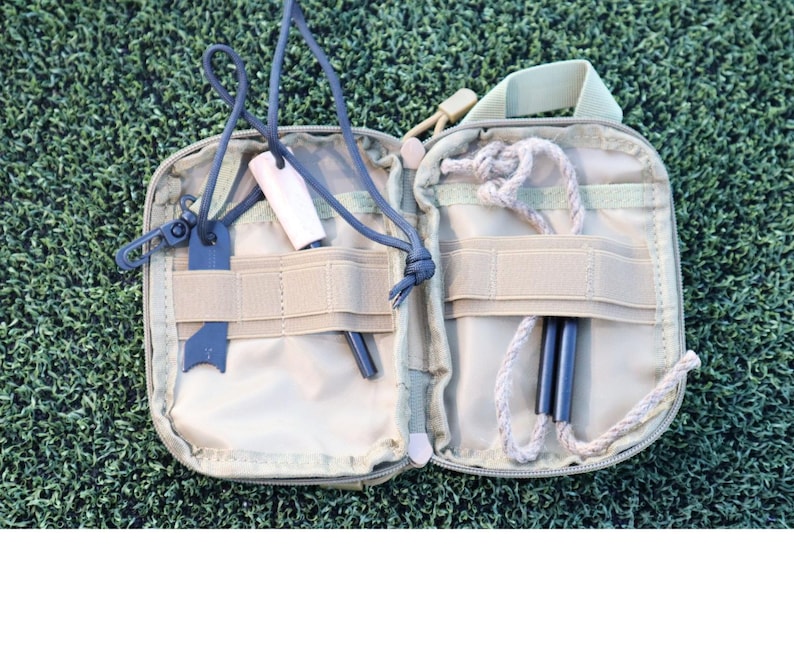 K&ouml;nnte beinhalten: Ge&ouml;ffnete, beigefarbene Survival-Kit-Tasche mit verschiedenen Werkzeugen. Enth&auml;lt einen schwarzen Feuerstarter, zwei Ferro-St&auml;be, einen Holzanz&uuml;nder und eine Schnur. Die Tasche ist ge&ouml;ffnet und zeigt den Inhalt vor einem gr&uuml;nen Hintergrund.
