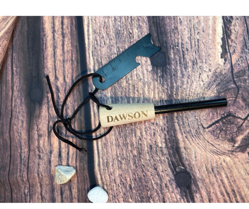 K&ouml;nnte beinhalten: Ein Feuerstarter-Set mit einem schwarzen Metalllineal, einem Holzgriff mit der Gravur "DAWSON" und einem schwarzen Stab. Das Set enth&auml;lt eine schwarze Schnur und befindet sich auf einem Holzgrund.