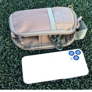 K&ouml;nnte beinhalten: Beige Tasche mit Mesh-Front und Rei&szlig;verschluss, Schl&uuml;sselanh&auml;nger und gr&uuml;nem Riemen. Ein wei&szlig;es Smartphone mit drei blauen Kameralinsen liegt daneben. Die Gegenst&auml;nde liegen auf einer gr&uuml;nen, strukturierten Oberfl&auml;che.