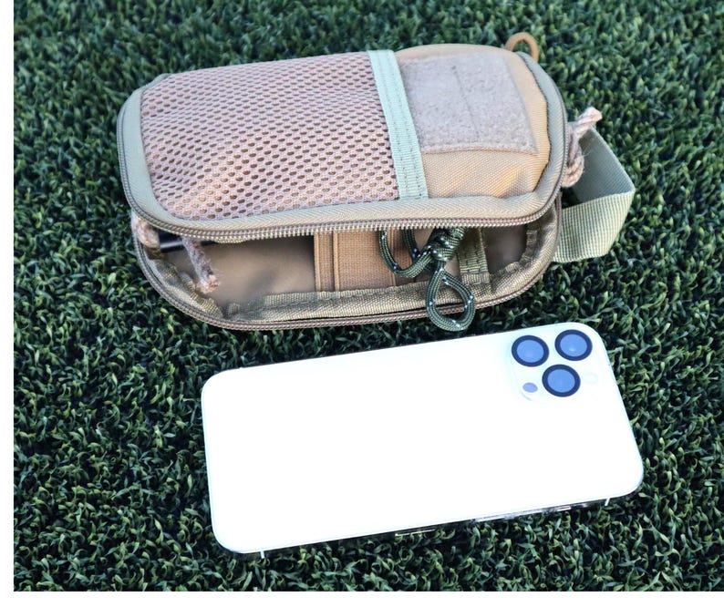 K&ouml;nnte beinhalten: Beige Tasche mit Mesh-Front und Rei&szlig;verschluss, Schl&uuml;sselanh&auml;nger und gr&uuml;nem Riemen. Ein wei&szlig;es Smartphone mit drei blauen Kameralinsen liegt daneben. Die Gegenst&auml;nde liegen auf einer gr&uuml;nen, strukturierten Oberfl&auml;che.