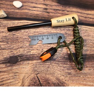 K&ouml;nnte beinhalten: Ein Feuerstarter-Set mit einem Holzgriff mit der Gravur "Stay Lit", einem schwarzen Metallz&uuml;nder, einem kleinen Lineal mit Millimeterangaben und einem gr&uuml;n-schwarzen Paracord-Lanyard. Das Set enth&auml;lt ein orange-schwarzes Werkzeug.