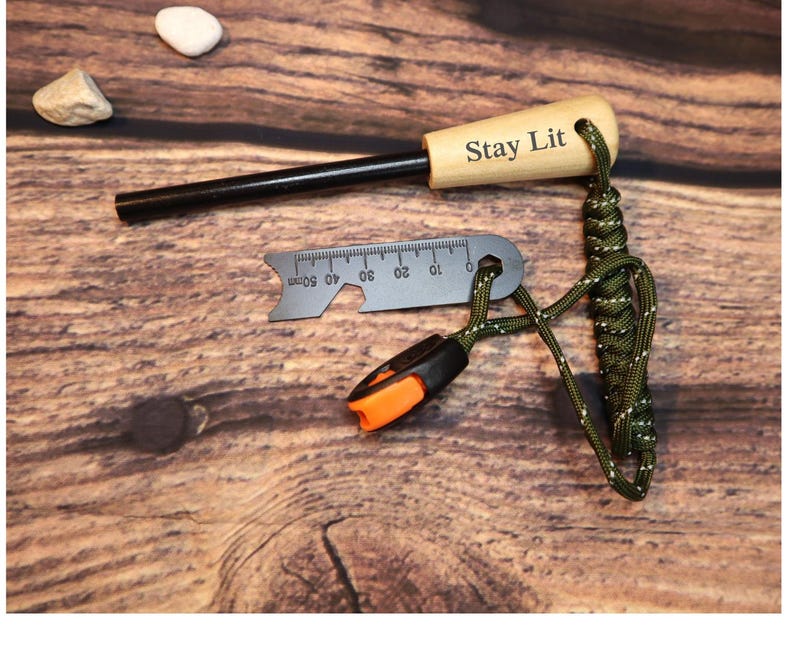 K&ouml;nnte beinhalten: Ein Feuerstarter-Set mit einem Holzgriff mit der Gravur "Stay Lit", einem schwarzen Metallz&uuml;nder, einem kleinen Lineal mit Millimeterangaben und einem gr&uuml;n-schwarzen Paracord-Lanyard. Das Set enth&auml;lt ein orange-schwarzes Werkzeug.