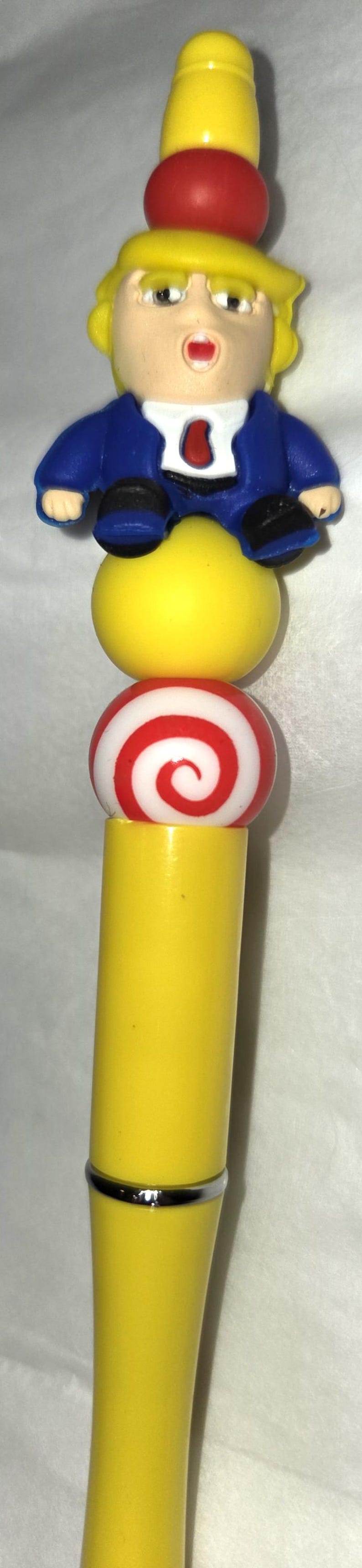 May include: A novelty pen featuring a cartoon figure with yellow hair and a blue suit. The pen has a yellow barrel, a red and white swirled ball, and a yellow top. The figure has a red tie.