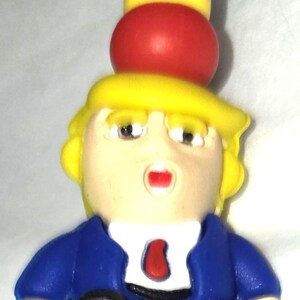 May include: A novelty figurine featuring a caricature with yellow hair, a blue suit, and a red tie. The figure has a red sphere and a yellow cylinder stacked on its head. The base is yellow, and the figure has a surprised expression.