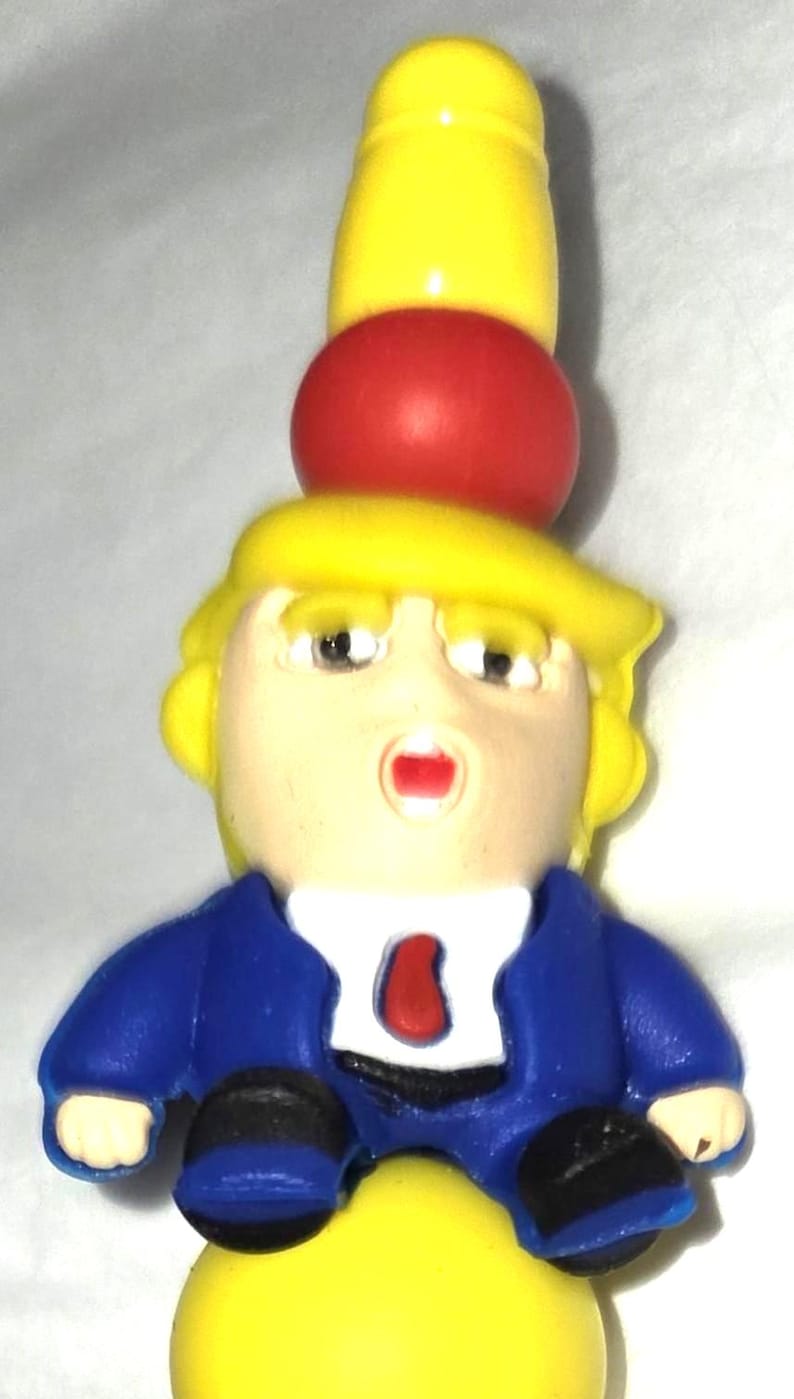 May include: A novelty figurine featuring a caricature with yellow hair, a blue suit, and a red tie. The figure has a red sphere and a yellow cylinder stacked on its head. The base is yellow, and the figure has a surprised expression.