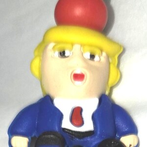 May include: A novelty figurine featuring a caricature with yellow hair, a blue suit, and a red tie. The figurine has a surprised expression and is perched atop a yellow and red base. The overall design is cartoonish.
