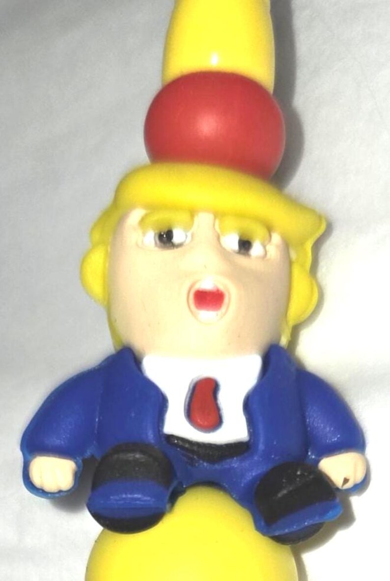 May include: A novelty figurine featuring a caricature with yellow hair, a blue suit, and a red tie. The figurine has a surprised expression and is perched atop a yellow and red base. The overall design is cartoonish.