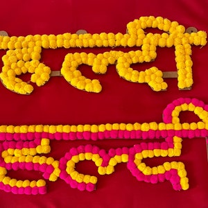 May include: Two words written in yellow and pink pom-poms on a red background. The words are "Happy" and "Birthday".