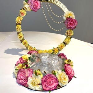 May include: A decorative floral arrangement featuring a gold-colored circular frame adorned with pink and yellow flowers, and faux pearls. The base is a mirrored surface with more flowers. The arrangement is likely for a wedding or special event.