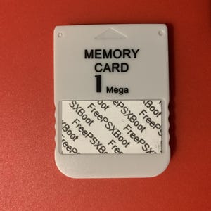 May include: A white memory card with the text "MEMORY CARD 1 Mega" printed in black. A sticker with the text "FreePSXBoot" is affixed to the bottom of the card. The card is on a red surface.