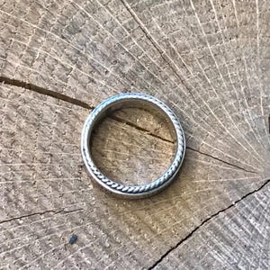 Op de afbeelding: Een zilveren ring met een touwachtig ontwerp langs de randen, geplaatst tegen een rustieke houten achtergrond. Het ontwerp van de ring is eenvoudig maar elegant en toont de fijne details.