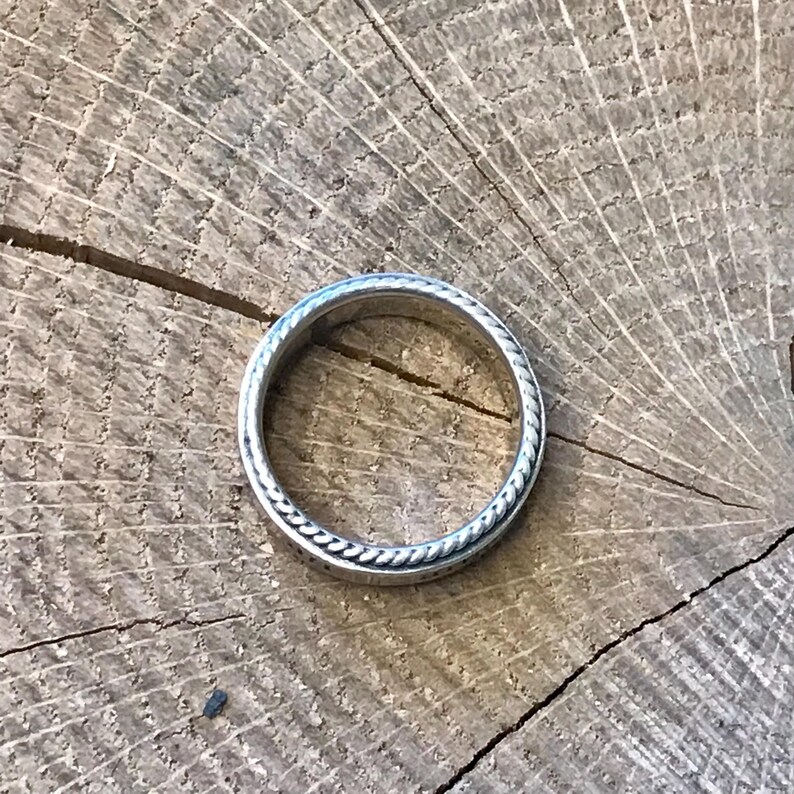 Op de afbeelding: Een zilveren ring met een touwachtig ontwerp langs de randen, geplaatst tegen een rustieke houten achtergrond. Het ontwerp van de ring is eenvoudig maar elegant en toont de fijne details.