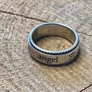 Op de afbeelding: Zilverkleurige ring met het woord "angel" gegraveerd op de band. De ring heeft een touwachtig ontwerp rond de randen en een klein engelsymbool. Geplaatst op een houten achtergrond.