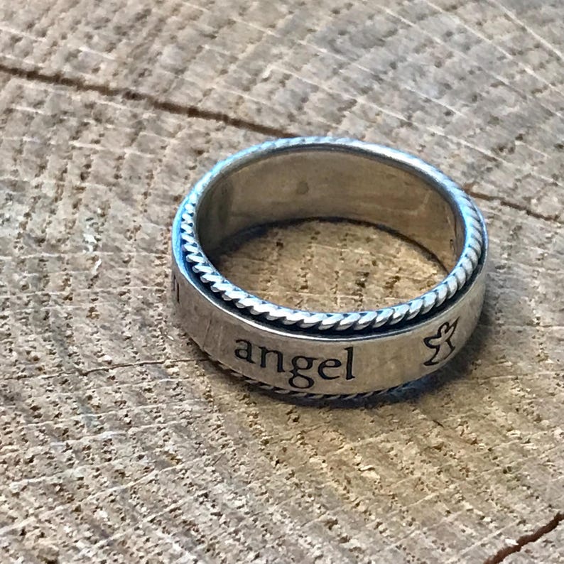 Op de afbeelding: Zilverkleurige ring met het woord "angel" gegraveerd op de band. De ring heeft een touwachtig ontwerp rond de randen en een klein engelsymbool. Geplaatst op een houten achtergrond.